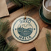 A decorative ornament with the text "GOZO MALTA" on it, featuring a cityscape design, is displayed on a wooden surface alongside a cup of coffee and some pine branches.