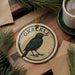 A round ornament with a black bird on it, sitting on a wooden surface, with a cup of coffee and pine branches nearby.