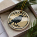 A round ornament with a black bird image and the word "GRACKLE" written on it, placed inside a box with pine branches.