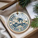 A decorative ornament with the text "GRAND CAYMAN ISLANDS" printed on it, surrounded by a palm tree and other tropical elements, placed in a box with pine branches.