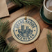 A decorative ornament with the text "GRAND RAPIDS MICHIGAN USA" printed on it, featuring a cityscape design. The ornament is hanging from a string and is placed on a wooden surface, surrounded by pine branches and a cup of coffee.