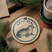 A ceramic ornament featuring a seal is displayed on a wooden surface, accompanied by a cup of coffee and pine branches.