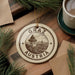 A circular ornament with the text "GRAZ AUSTRIA" and an illustration of a building, placed on a wooden surface next to a cup of coffee and pine branches.