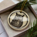 A ceramic ornament in the shape of an owl is displayed in a box, surrounded by pine branches.
