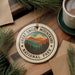 A circular ornament with the text "GREAT SMOK MOUNTAINS NATIONAL PARK" is displayed on a wooden surface, surrounded by pine branches and a cup of coffee.