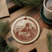 a round ornament with a castle on a hill, surrounded by pine branches and a cup of coffee on a wooden surface.