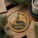 A circular ornament featuring a loon, a type of bird, is displayed on a wooden surface alongside a cup of coffee and some pine branches.
