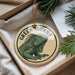 A circular ornament featuring a green iguana, with the text "GREEN IGuana" printed on it, is displayed in a box with pine branches.