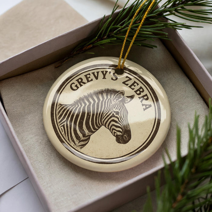 A circular ornament with a zebra's head image and the text "GREVY'S ZEBRA" on it, placed in a box with pine branches.