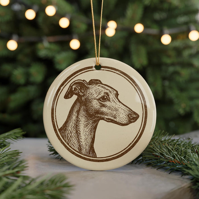 A round ornament featuring a greyhound dog's head is displayed against a blurred background of Christmas lights.