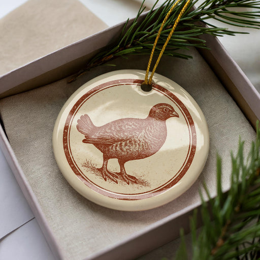 A ceramic ornament in the shape of a bird, possibly a quail, is displayed in a box with pine branches.