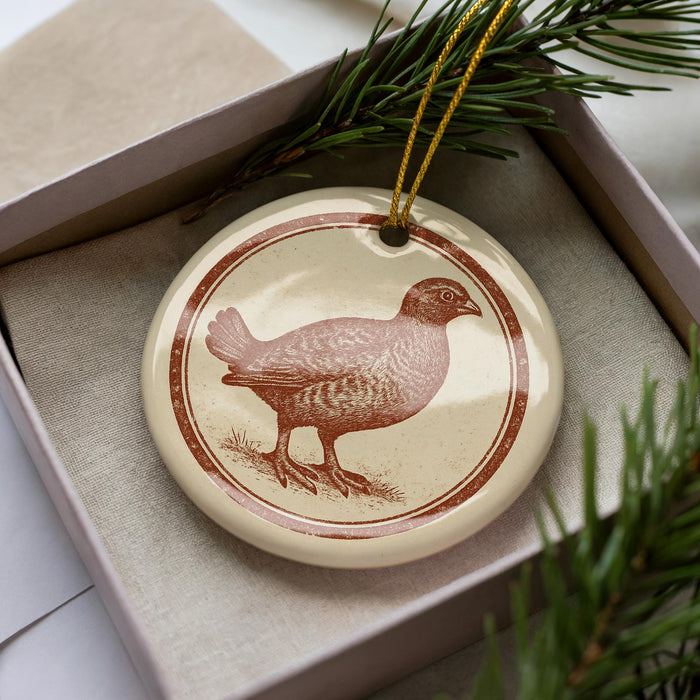 A ceramic ornament in the shape of a bird, possibly a quail, is displayed in a box with pine branches.