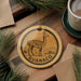 A decorative ornament featuring a llama, with the word "GUANACO" written on it, is displayed on a wooden surface alongside a cup of coffee and some pine branches.
