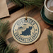 A circular ornament with the text "GUERNSEY CHANNEL ISLANDS" and a graphic of a map of the Channel Islands, hanging on a string. The ornament is surrounded by pine branches and a cup of coffee on a wooden surface.