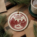 A decorative ornament with a red and white design, featuring mountains, a boat, and the text "GUILIN CHINA", is displayed on a wooden surface alongside a cup of coffee and some pine branches.