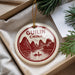 A decorative ornament with a red and white design, featuring mountains, a river, and a person on a boat, is displayed in a box with pine branches.