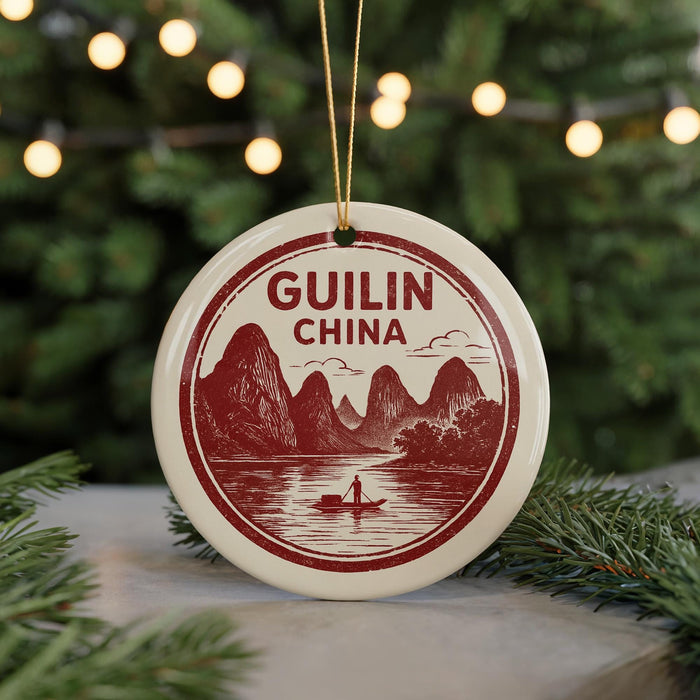 A circular ornament with a red and white design depicting mountains, a river, and a boat, hanging from a string.