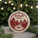 A circular ornament with a red and white design depicting mountains, a river, and a boat, hanging from a string.