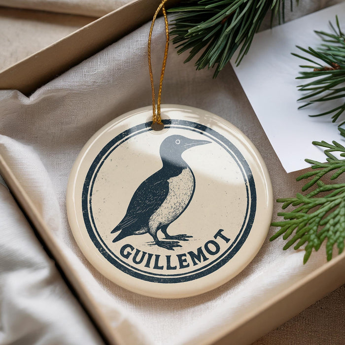 A decorative ornament featuring a black and white illustration of a bird, with the text "GUILLLEMOT" printed on it, is displayed in a box with pine branches.