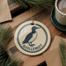 A decorative ornament featuring a black and white illustration of a bird, likely a goose, with the word "GUILLLEMOT" written on it, is displayed on a wooden surface alongside a cup of coffee and some pine branches.