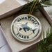 A circular ornament with a hammerhead shark design and the text "HAMMERSHEAD SHARK" printed on it, placed inside a box with pine branches.