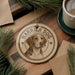 A circular ornament featuring a dog's head and the text "Harrier Dog" is displayed on a wooden surface, surrounded by pine branches and a cup of coffee.