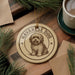 A round ornament featuring a dog illustration, with the text "HAVANESE DOG" around the edge. The ornament is placed on a wooden surface, with pine branches and a cup of coffee nearby.