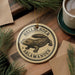 A circular ornament featuring a salamander, with the text "HELLBENDER SALAMANDER" printed on it, is displayed on a wooden surface alongside a cup of coffee and some pine branches.