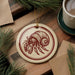 a round ornament with a red and white design depicting a hermit crab, placed on a wooden surface next to a cup of coffee and some pine branches.