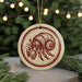 A round ornament featuring a red and white illustration of a hermit crab is displayed against a blurred background of a Christmas tree with twinkling lights.