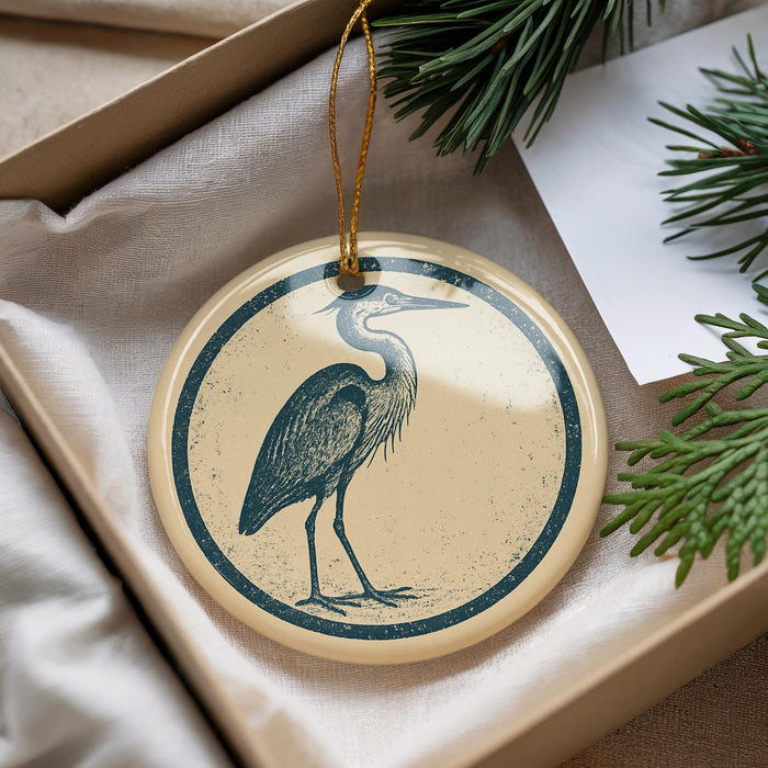 A circular ornament featuring a blue heron is displayed in a box, surrounded by pine branches and a white card.