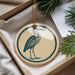 A circular ornament featuring a blue heron is displayed in a box, surrounded by pine branches and a white card.