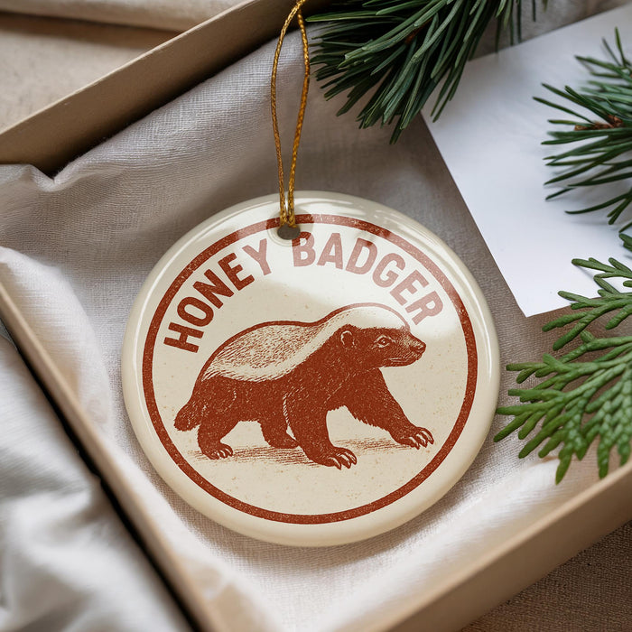 A round ornament with the text "HONEY BADGER" and an illustration of a badger is displayed in a box with pine branches.
