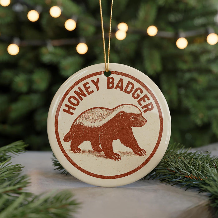A round ornament with the text "HONEY BADGER" and an illustration of a badger is displayed in front of a Christmas tree with twinkling lights.