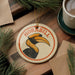 A hornbill ornament with the word "HORNBILL" is displayed on a wooden surface, surrounded by pine branches and a cup of coffee.