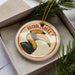 A round ornament with a hornbill illustration on it, placed in a box with pine branches.