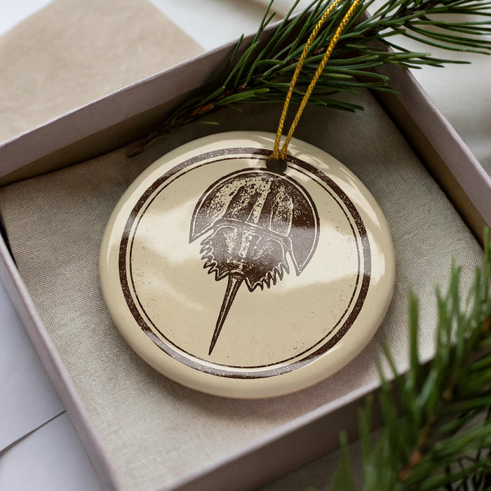 A circular ornament with a brown design featuring a stylized image of a sea creature, possibly a crab or lobster, is displayed in a box with pine branches.