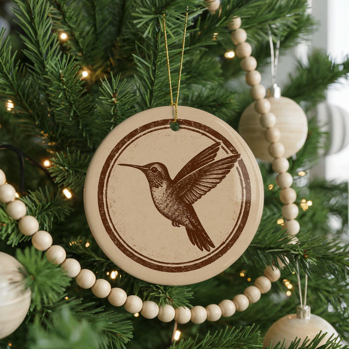 A round ornament with a brown bird design hangs from a Christmas tree, surrounded by a string of beads and other ornaments.