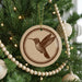 A round ornament with a brown bird design hangs from a Christmas tree, surrounded by a string of beads and other ornaments.