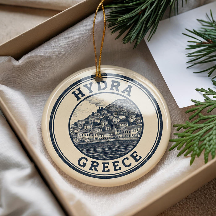 A round ornament with the text "HYDRA GREECE" and an illustration of a town, placed on a beige surface next to some pine branches.