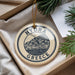 A round ornament with the text "HYDRA GREECE" and an illustration of a town, placed on a beige surface next to some pine branches.