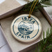 A circular ornament with the text "IBiza Spain" printed on it, placed inside a box with pine branches.