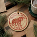 A round ornament featuring a tiger illustration, with the text "INDOCOCHINESE TIGER" printed on it, is displayed on a wooden surface alongside a cup of coffee and some pine branches.