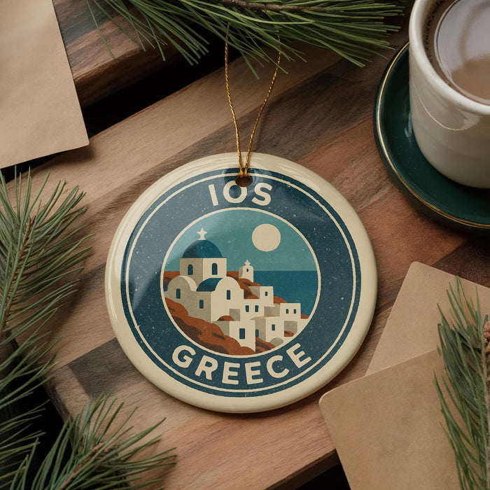 A circular ornament with a design of a Greek island and the text "10OS GREECE" is displayed on a wooden surface, surrounded by pine branches and a cup of coffee.