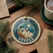 A circular ornament with a design of a Greek island and the text "10OS GREECE" is displayed on a wooden surface, surrounded by pine branches and a cup of coffee.