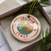 A decorative ornament in a box, featuring a sailboat and the text "ISCHIA ITALY" on a circular design.