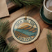 A circular ornament with the text "ISLA DEL SOL BOLIVIA" is displayed on a wooden surface, surrounded by pine branches and a cup of coffee.