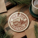 A circular ornament with the text "ISLE OF SKYE SCOTLAND" printed on it, surrounded by pine branches and a cup of coffee on a wooden surface.