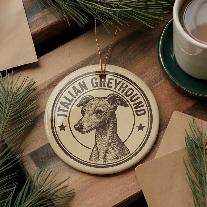 A circular ornament featuring an illustration of a greyhound dog, with the text "ITALIAN GREYHOUND" around the edge. The ornament is placed on a wooden surface, surrounded by pine branches and a cup of coffee.