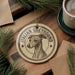 A circular ornament featuring an illustration of a greyhound dog, with the text "ITALIAN GREYHOUND" around the edge. The ornament is placed on a wooden surface, surrounded by pine branches and a cup of coffee.